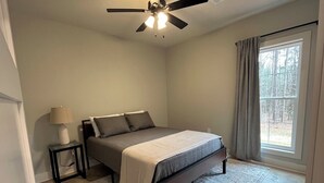 A bedroom with a bed, a nightstand, a lamp, a ceiling fan, and a window with curtains. - The Gray Cottage on Brownlee! (Calhoun)