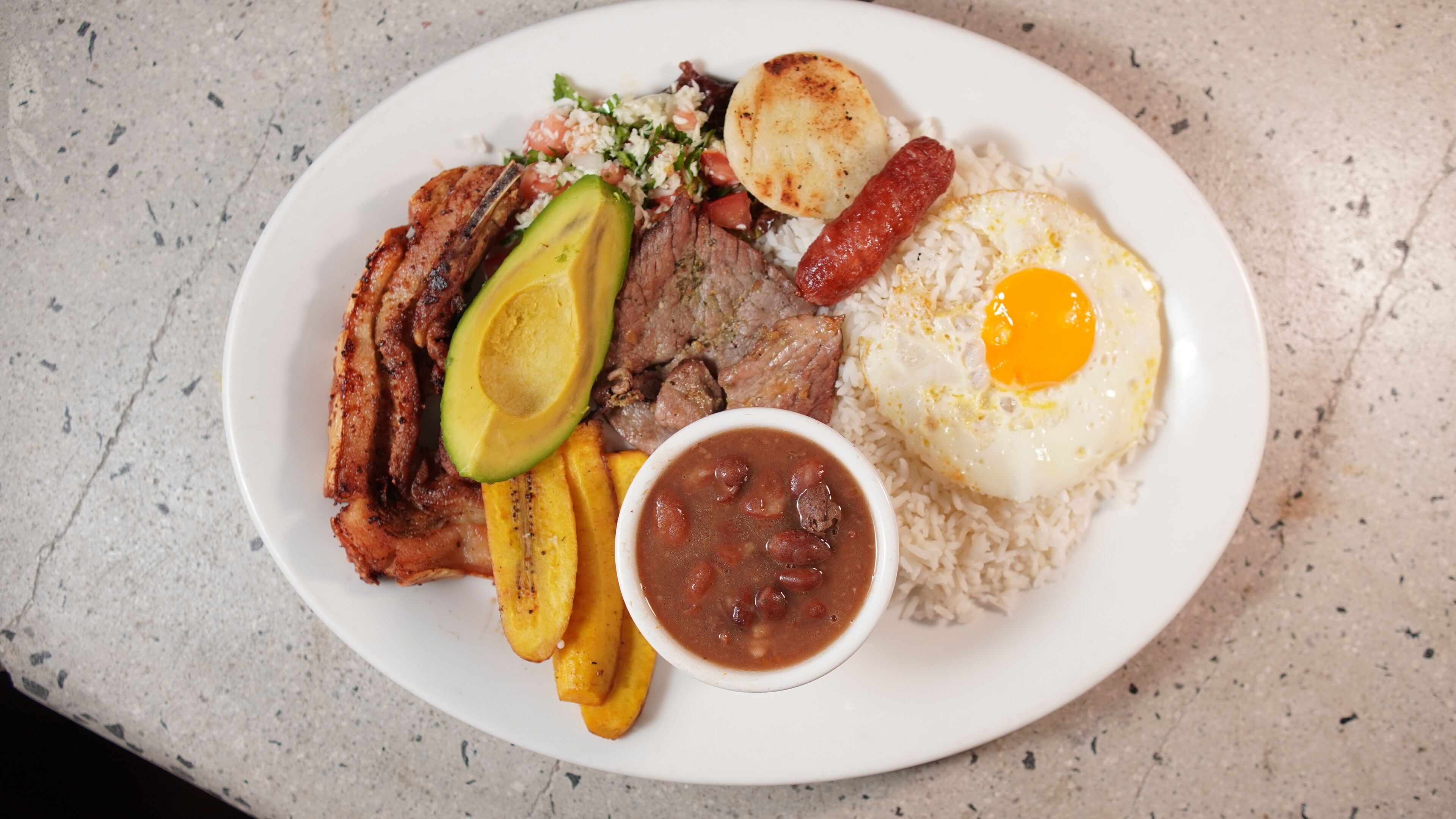 A plate of food with rice, a fried egg, avocado, beans, plantains, and a sausage.