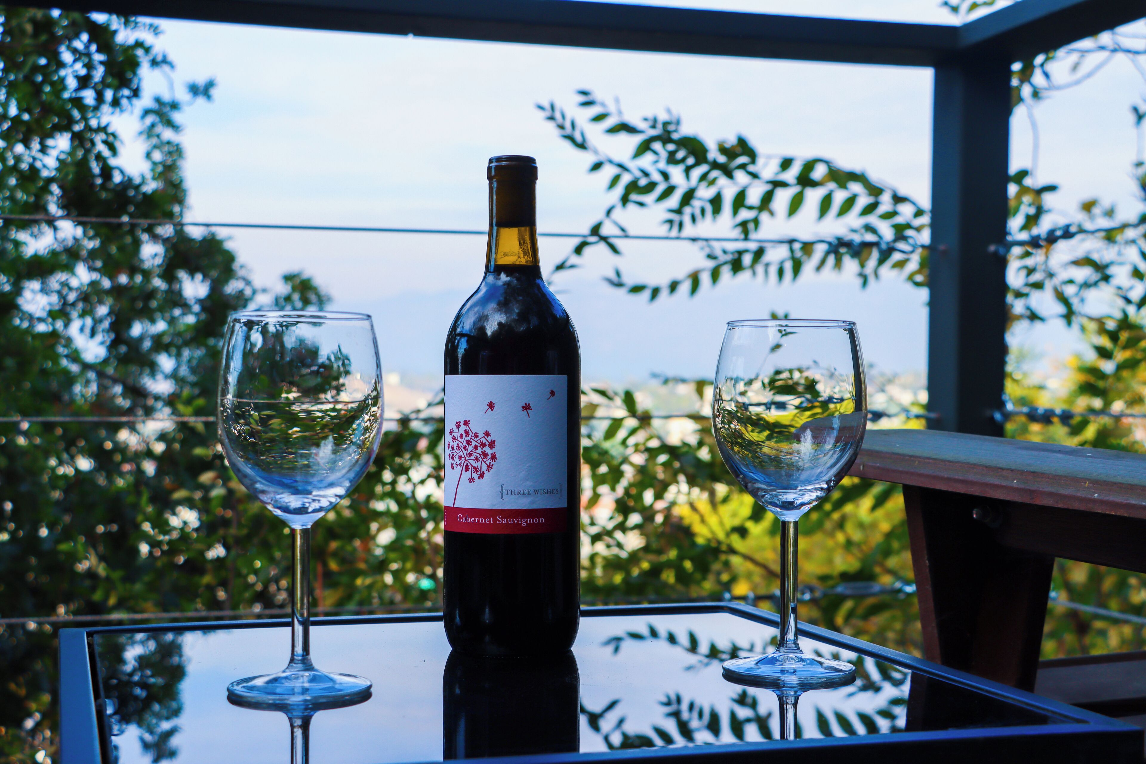 Eine Flasche Cabernet Sauvignon Wein auf einem Glastisch, zusammen mit zwei leeren Weingläsern, vor einer Kulisse aus Bäumen und blauem Himmel.
