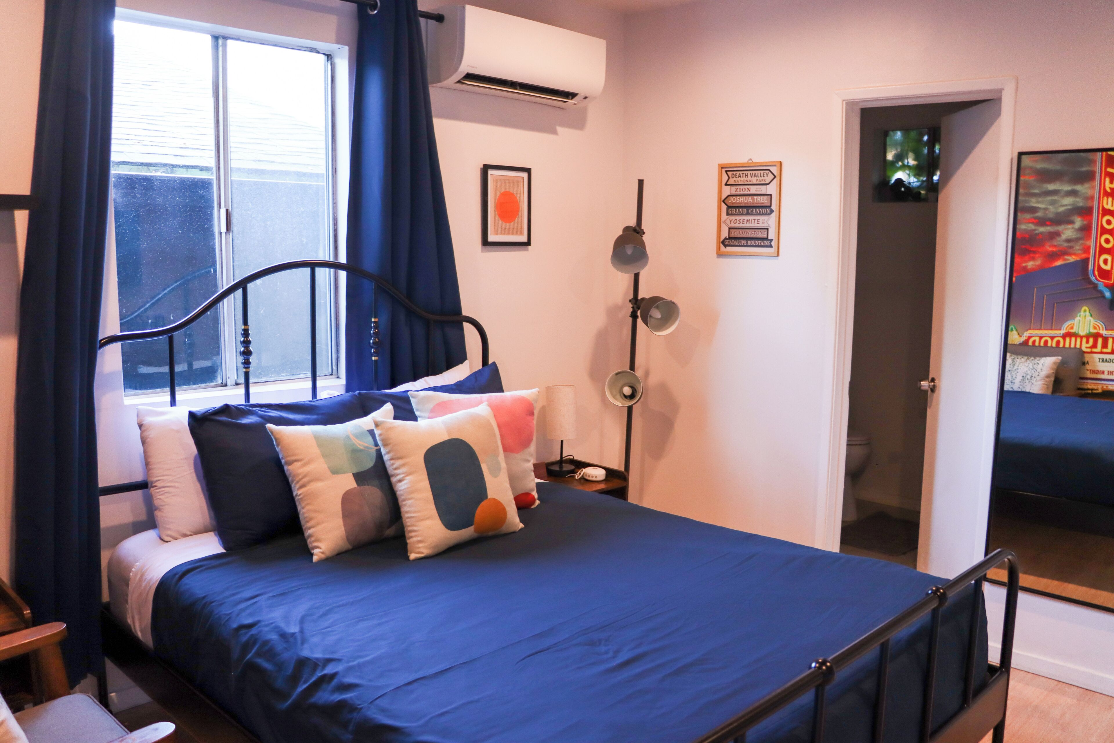 Een slaapkamer met een bed, een nachtkastje, een lamp, een raam met gordijnen, een airconditioner, een deur en een poster aan de muur.