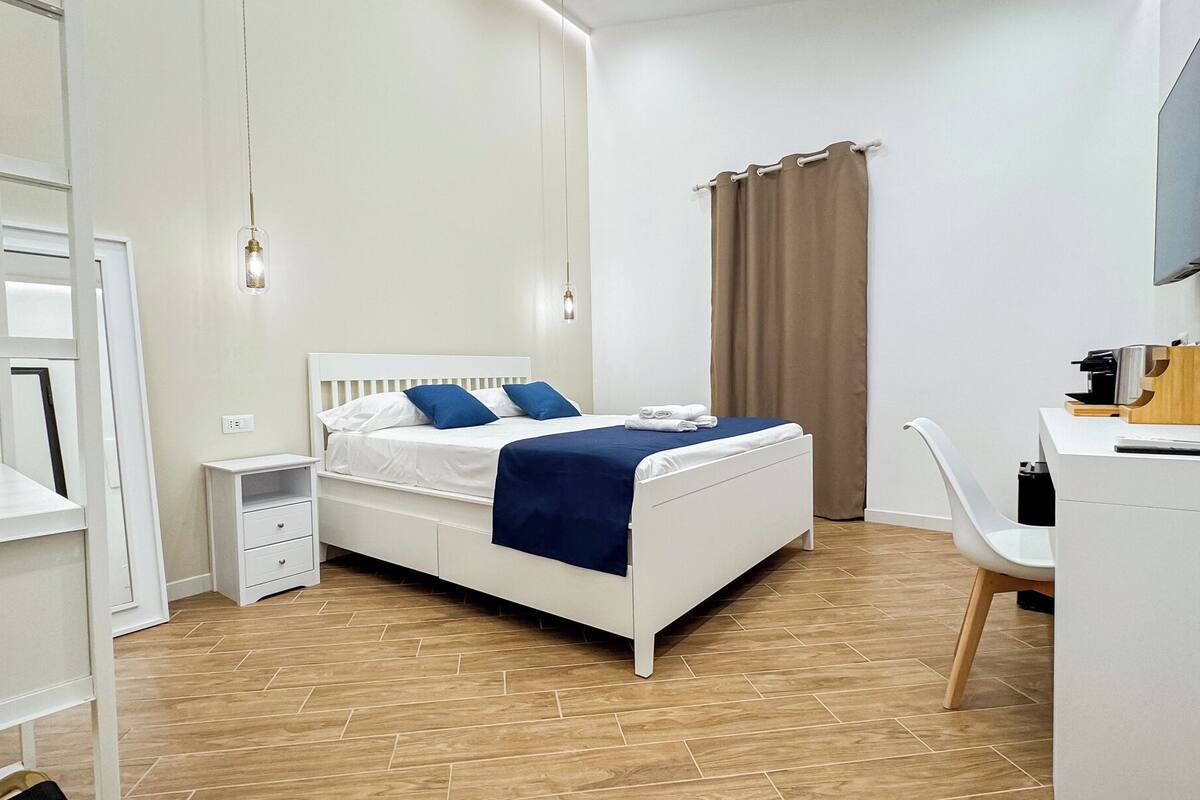 A modern bedroom with a white bed, blue bedding, a desk with a chair, and a TV mounted on the wall.