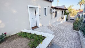 Un sendero estrecho que lleva a un edificio con una puerta blanca, unidades de aire acondicionado y una ventana pequeña.