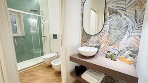 Un baño moderno con ducha de vidrio, un lavamanos blanco con un recipiente y un muro con estampado floral.