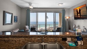 A hotel room with a kitchen area, a large window with a view of the ocean, a ceiling fan, and a flat-screen TV mounted on the wall. - Ariel Dunes II 2105 (Miramar Beach)