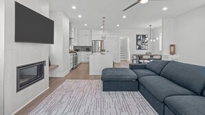 A modern living room with a grey sectional sofa, a fireplace, and a kitchen in the background. - Spacious 4 Bedroom House in Charming Downtown Raleigh with Amazing Views! (Raleigh)