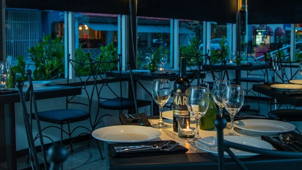 A restaurant setting with tables set for dining, wine glasses, and bottles, with a view of the outside.