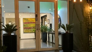 A hotel entrance with glass doors, potted plants, a colorful abstract painting, and a visible interior space with a table and chairs.