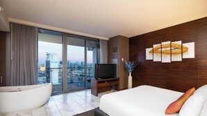 Una habitación de hotel moderna con una cama grande, una bañera, televisión y vistas a la ciudad a través de amplios ventanales.