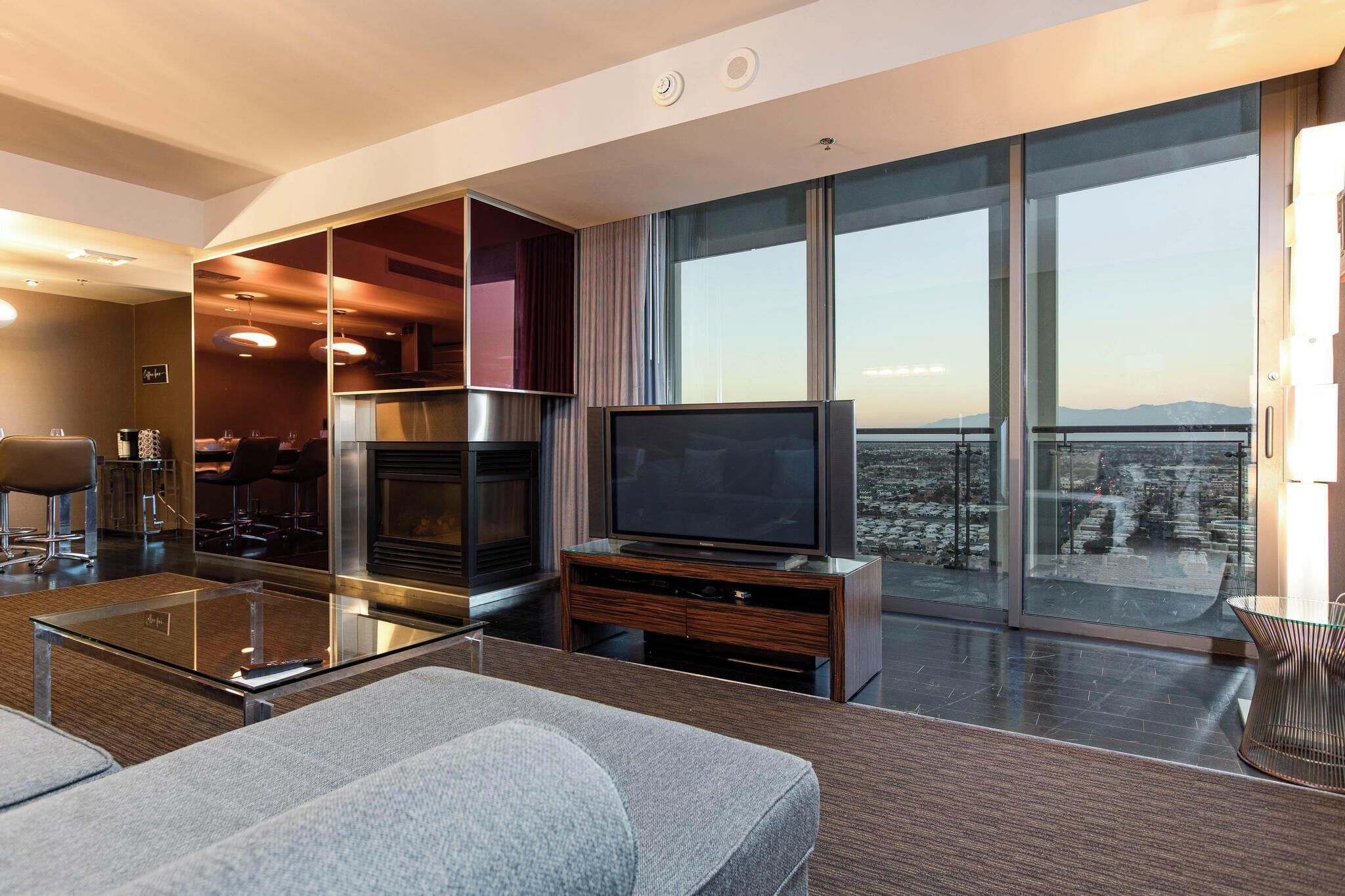 Una habitación de hotel moderna con un televisor grande, una mesa de centro de vidrio, un sofá y un balcón con vistas a la ciudad.