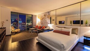 Habitación de hotel moderna con una cama grande, un sofá, un escritorio y vistas a la ciudad a través de ventanales de pared a techo.