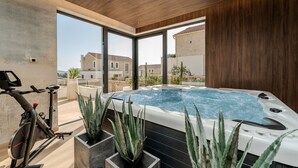 A modern indoor pool area with a stationary bike, large windows, and a view of a stone building. - Luxury Villa Waterlily with Pool (Vodice)