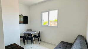 A compact living space with a grey sofa, a dining table with chairs, and a TV mounted on the wall. - CASA DAS FLORES (Praia Grande)