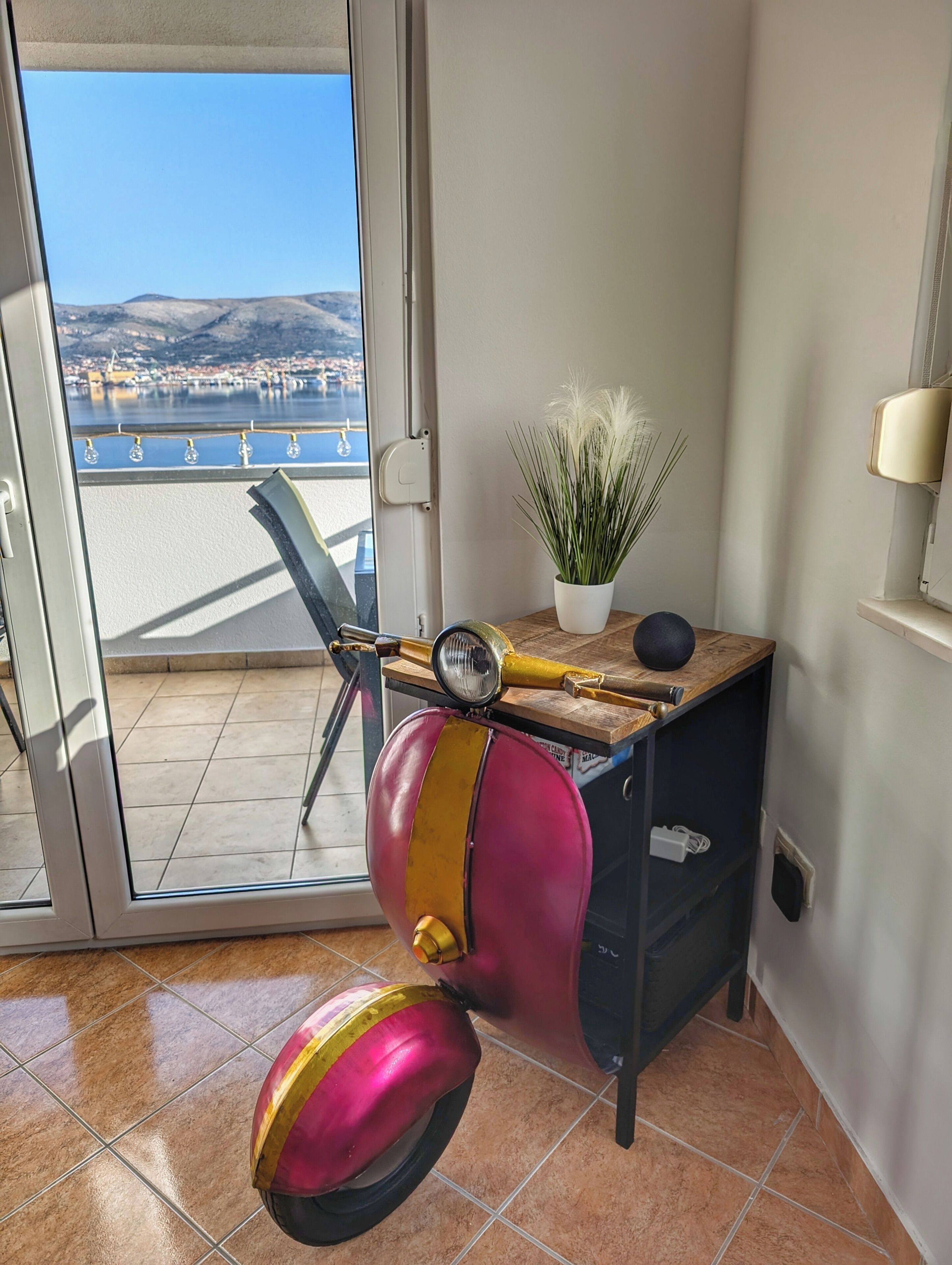 A room with a view of a body of water and mountains, a potted plant, a vintage radio, and a scooter.