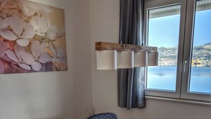 Un coin repas avec une table blanche, des chaises noires, un saladier rempli de fruits et une vue sur une étendue d’eau par la fenêtre.