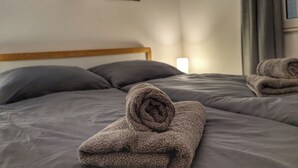 Un lit bien fait, avec une literie grise et une serviette enroulée posée sur le dessus. Trois tableaux représentant des motifs floraux sont accrochés au mur, au-dessus du lit.