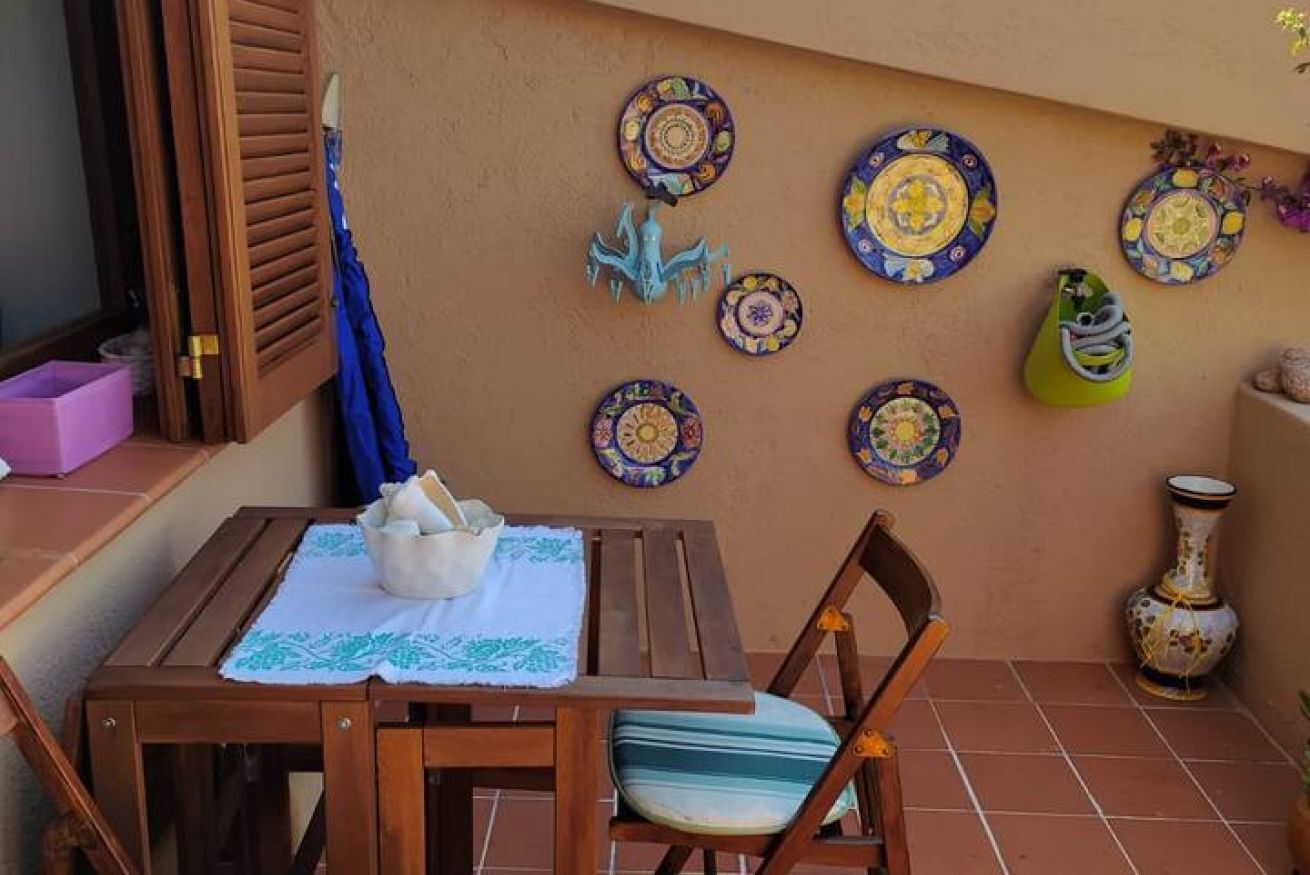 Une table en bois recouverte d’une nappe blanche, une chaise avec un coussin à rayures et un parasol bleu appuyé contre un mur orné d’assiettes décoratives.
