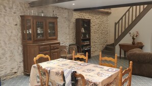 Ein Essbereich mit Tisch für vier Personen, umgeben von Holzstühlen. Links befindet sich ein Schrank mit Glastüren, im Hintergrund eine Bücherwand. Eine Holztreppe führt zu einer oberen Ebene.