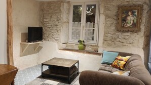 Ein gemütliches Wohnzimmer mit Steinwand, einem Holztisch, einem braunen Sofa mit bunten Kissen, einem Fernseher auf einem Regal und einer Vase mit Blumen auf der Fensterbank.