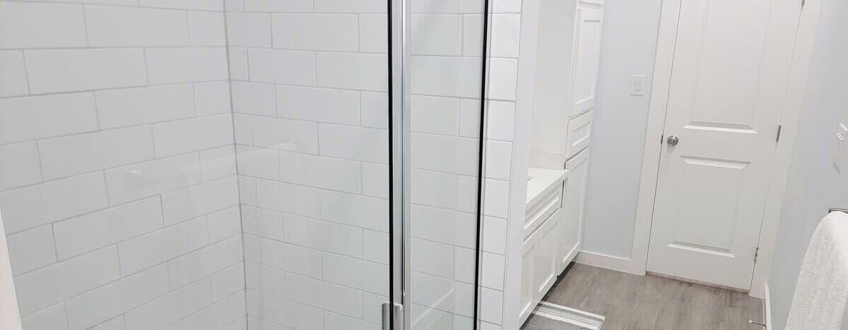 Une salle de bain avec des murs recouverts de carreaux blancs, une douche en verre et une porte blanche.