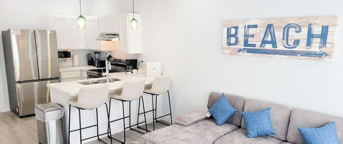 A modern kitchen with a central island, white bar stools, and a stainless steel refrigerator.