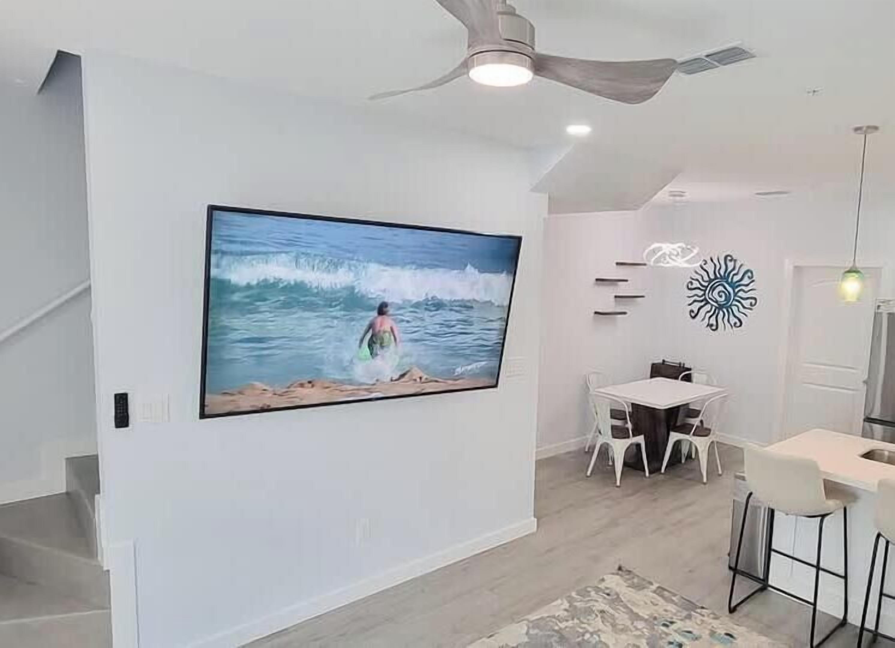 Una sala de estar moderna con un televisor de pantalla plana grande montado en la pared, un ventilador de techo, una zona de comedor con mesa y sillas, y una cocina con fregadero y taburetes de bar.