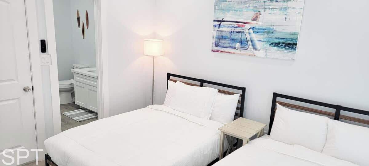 A hotel room with two beds, a bedside table, a lamp, and a painting on the wall.