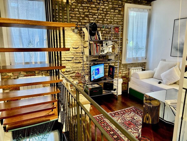 Un salon moderne avec un escalier en bois, un canapé blanc, un tapis à motifs et une bibliothèque adossée à un mur de briques.