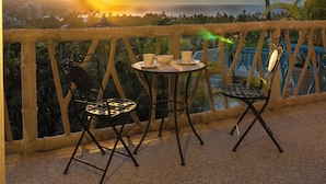 Un balcon avec une table et des chaises, offrant une vue sur un coucher de soleil, avec des palmiers et une silhouette de ville en arrière-plan.