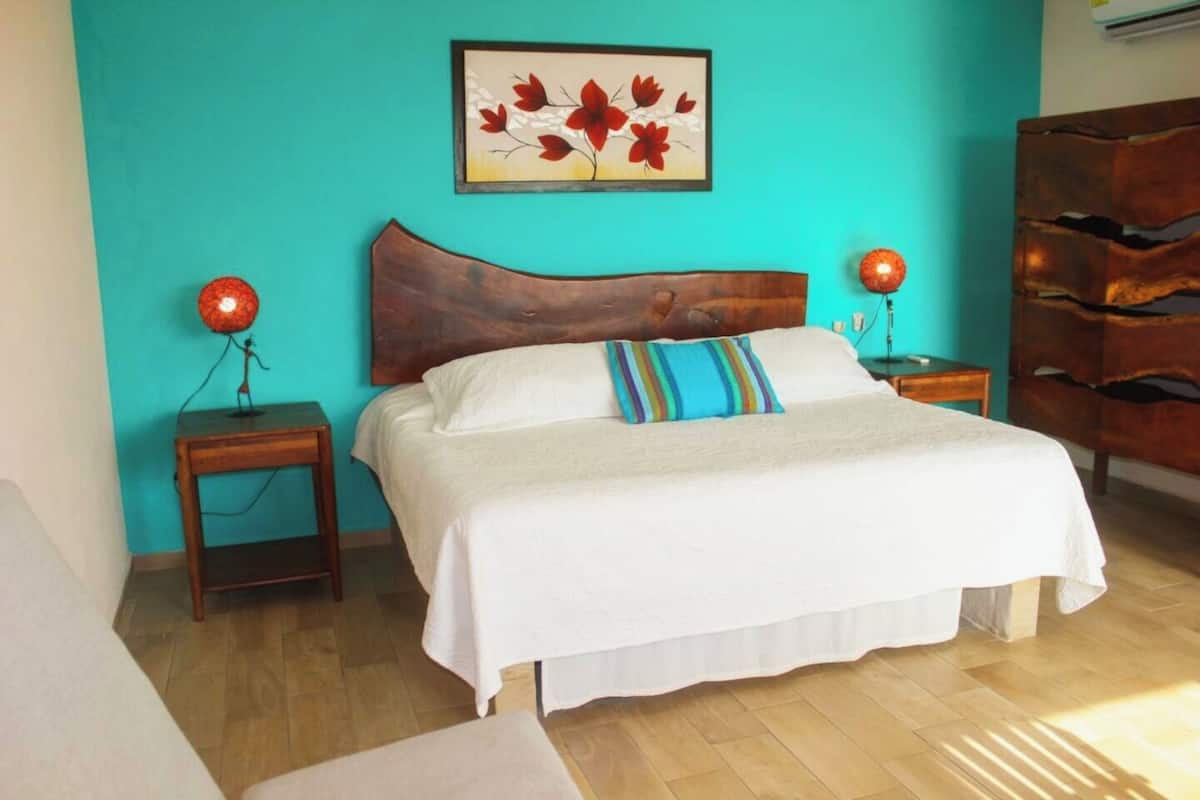 Une chambre à coucher avec un grand lit, deux tables de chevet, une tête de lit en bois, un tableau accroché au mur et une commode en bois.