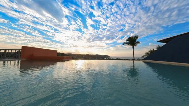 Une piscine avec vue sur le coucher de soleil, des palmiers et une côte lointaine.