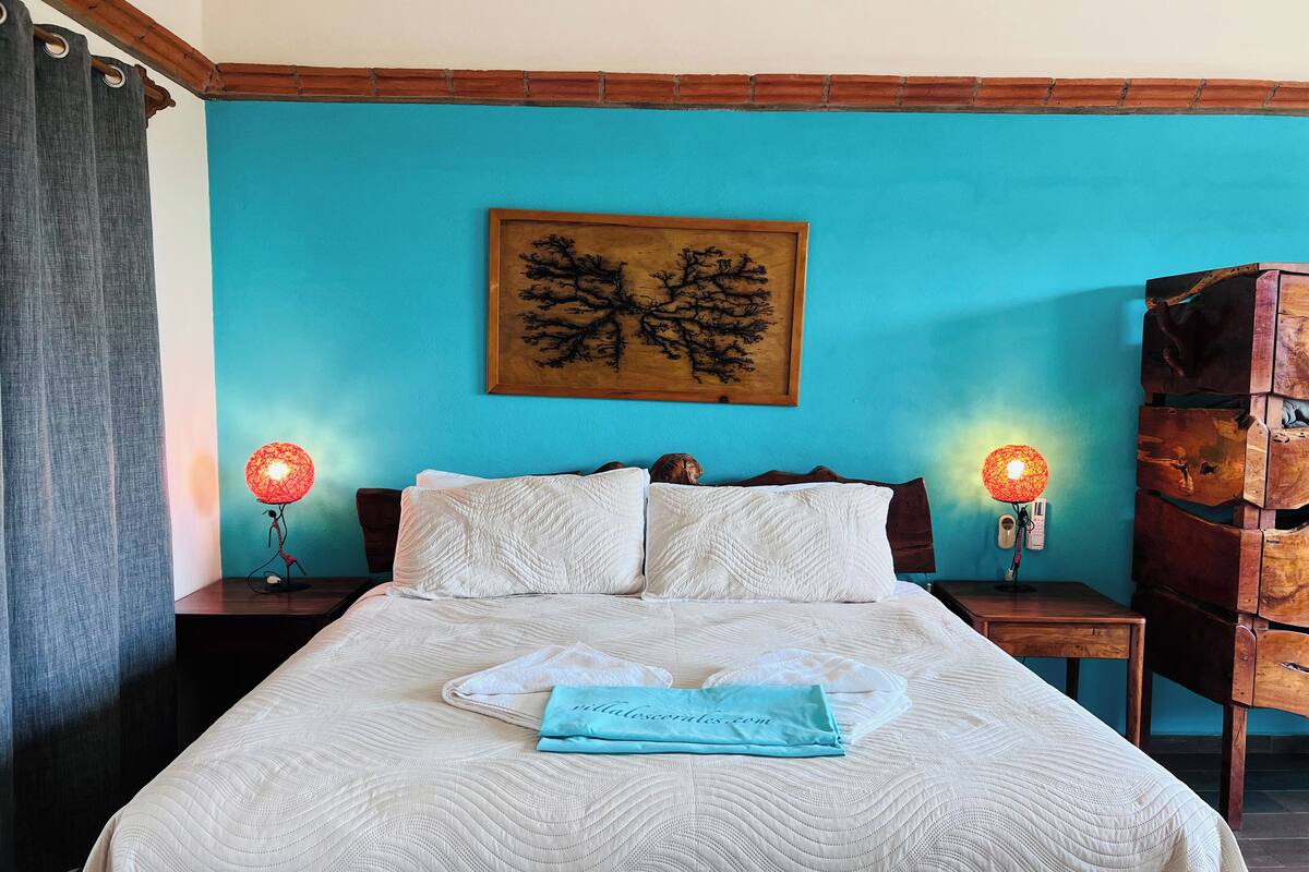 Un lit bien fait, recouvert d’une couette blanche, avec deux oreillers et une serviette pliée sur laquelle figure l’adresse d’un site web. On y trouve deux lampes de chevet à abat-jour rouge, une tête de lit en bois et un tableau représentant un arbre, accroché au mur.