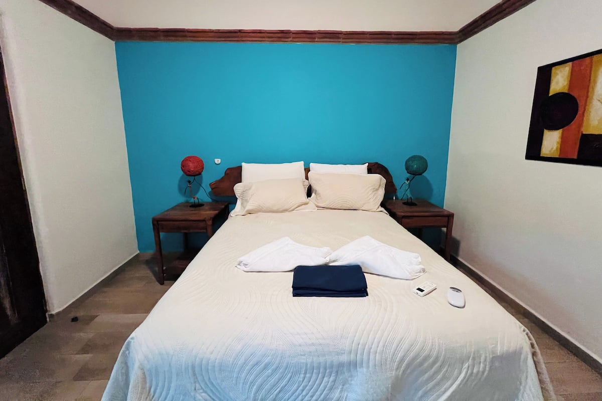 Un lit bien fait, avec des oreillers et une serviette pliée, une table de chevet avec une lampe et un tableau accroché au mur.
