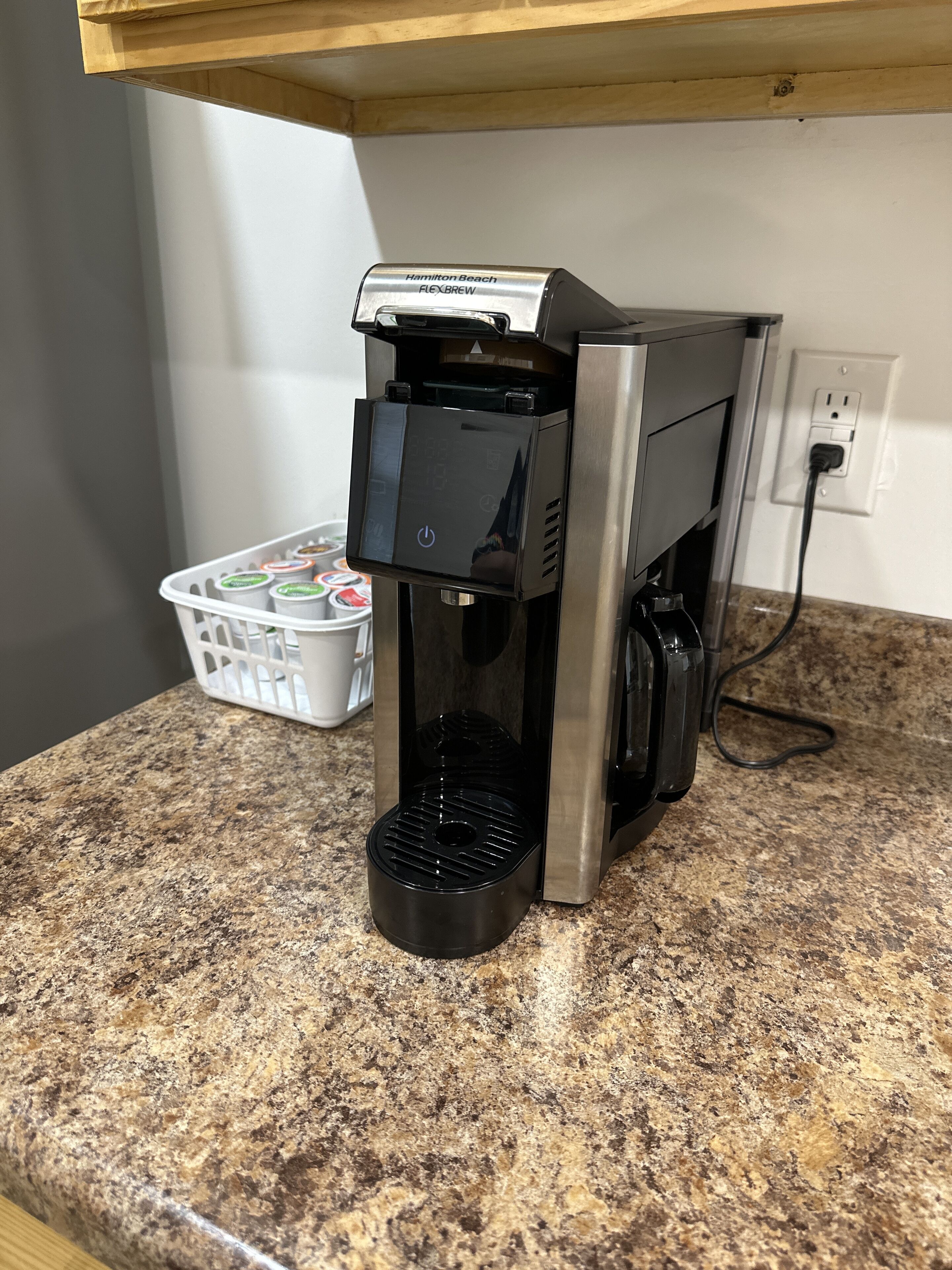 Una cafetera Hamilton Beach FlexBrew sobre un taulell de granat, amb una cistella blanca de càpsules de cafè al fons.