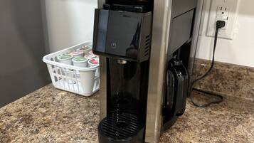 Una cafetera Hamilton Beach FlexBrew sobre un taulell de granat, amb una cistella blanca de càpsules de cafè al fons.