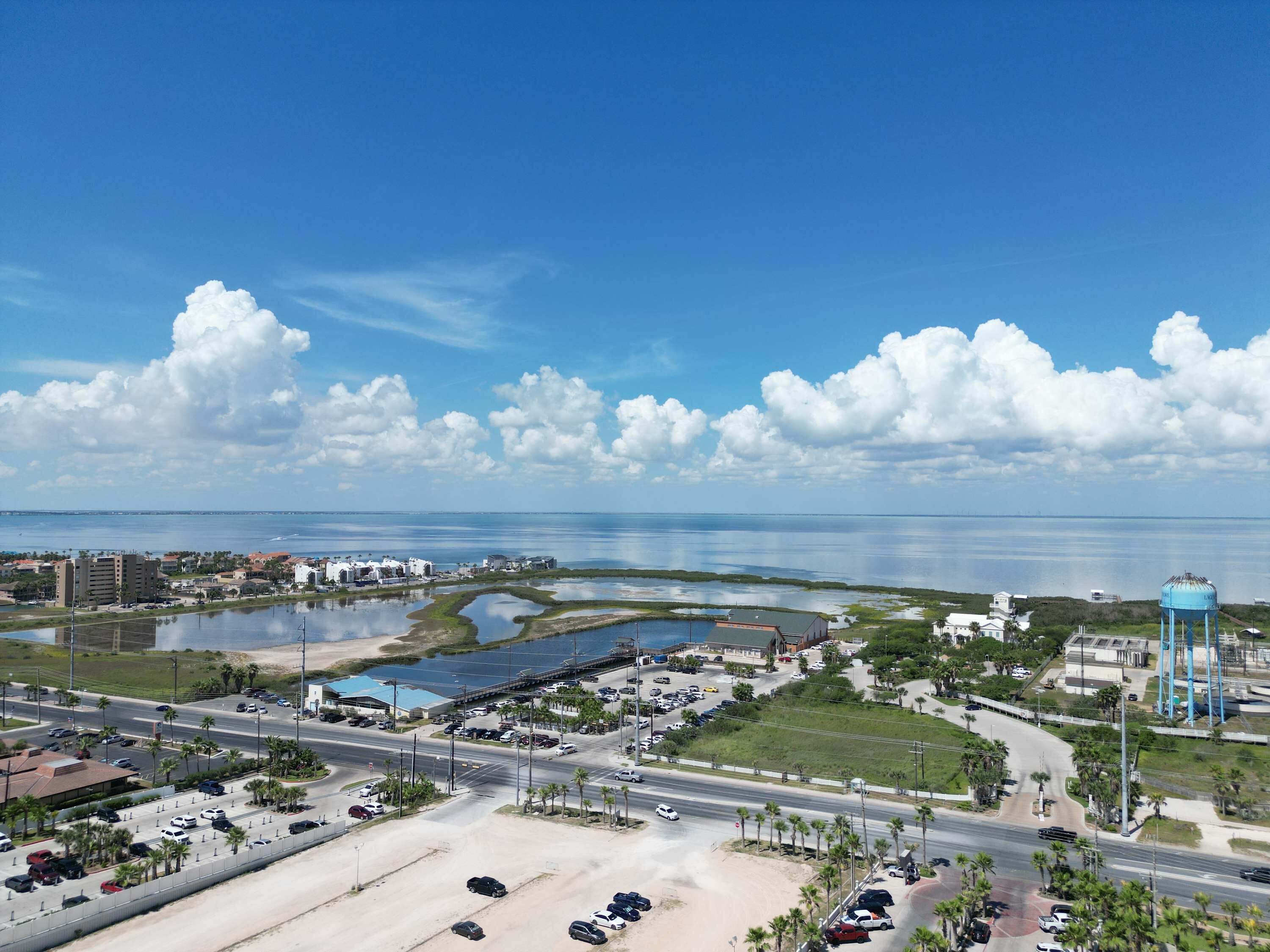A coastal cityscape with a prominent water tower, a marina, and a road leading to a parking area.