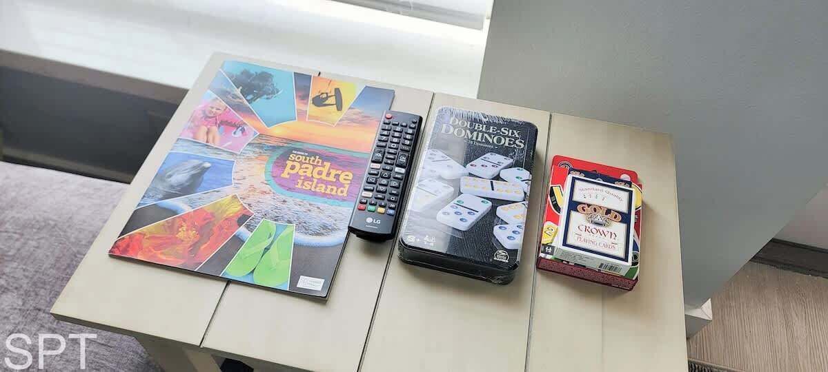 A table with a South Padre Island book, an LG remote, a Dominoes game, and a Gold Crown training card.