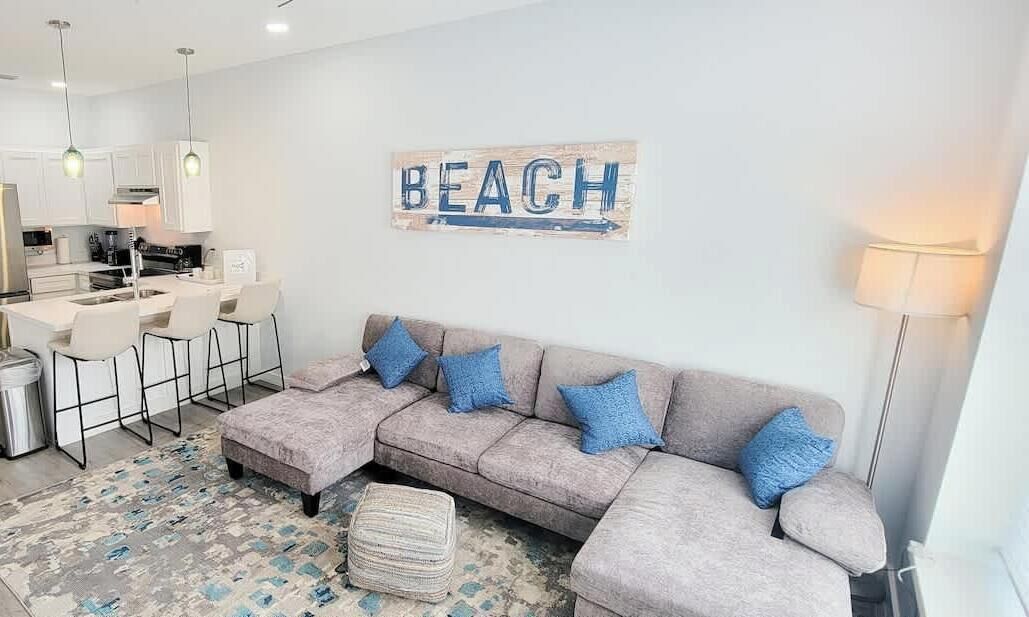 A modern living room with a grey sectional sofa, a coffee table, and a rug. There is a kitchen area with white cabinets and a dining table with chairs. A wall-mounted sign reads "BEACH".