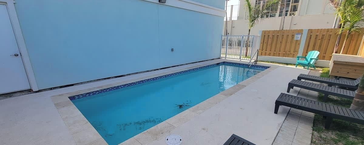 A rectangular swimming pool with a tiled edge, surrounded by a light-colored deck. There are lounge chairs and a green chair near the pool.