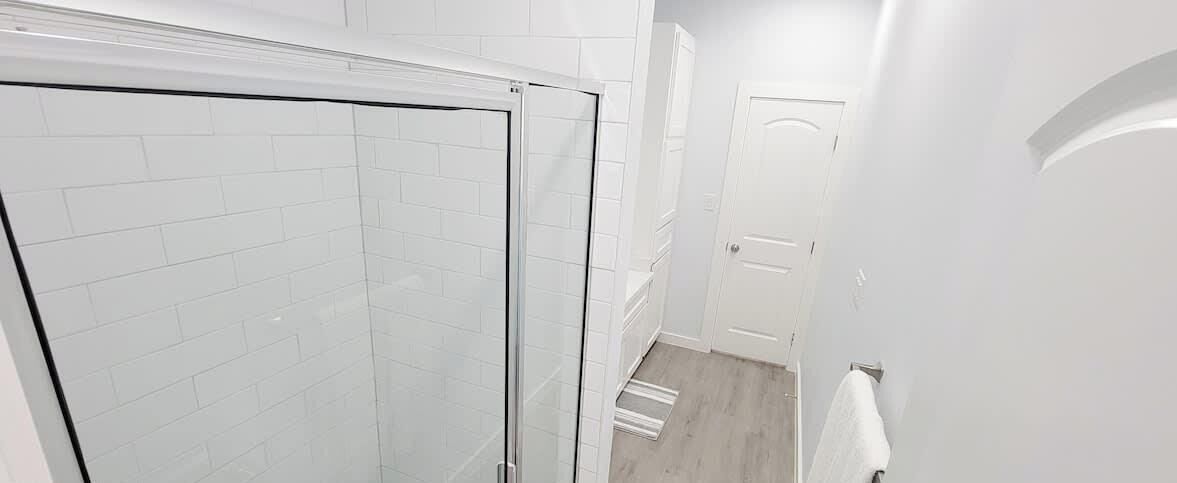 Baño con paredes de azulejos blancos, cabina de ducha de vidrio y suelo de madera.