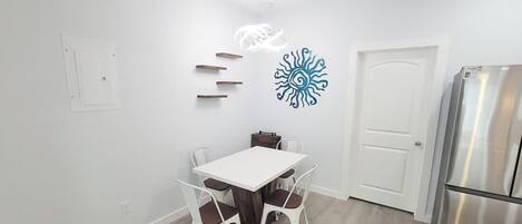 Una cocina con una mesa blanca, cuatro sillas, una puerta blanca y un refrigerador de acero inoxidable.