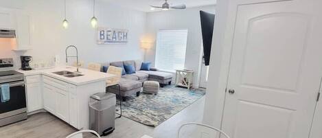 A modern kitchen with white cabinets, a built-in oven, and a sink. There is a dining area with a white table and chairs. A gray sofa and ottoman are in the living room, which has a patterned rug. A ceiling fan and a sign reading "BEACH" are visible.