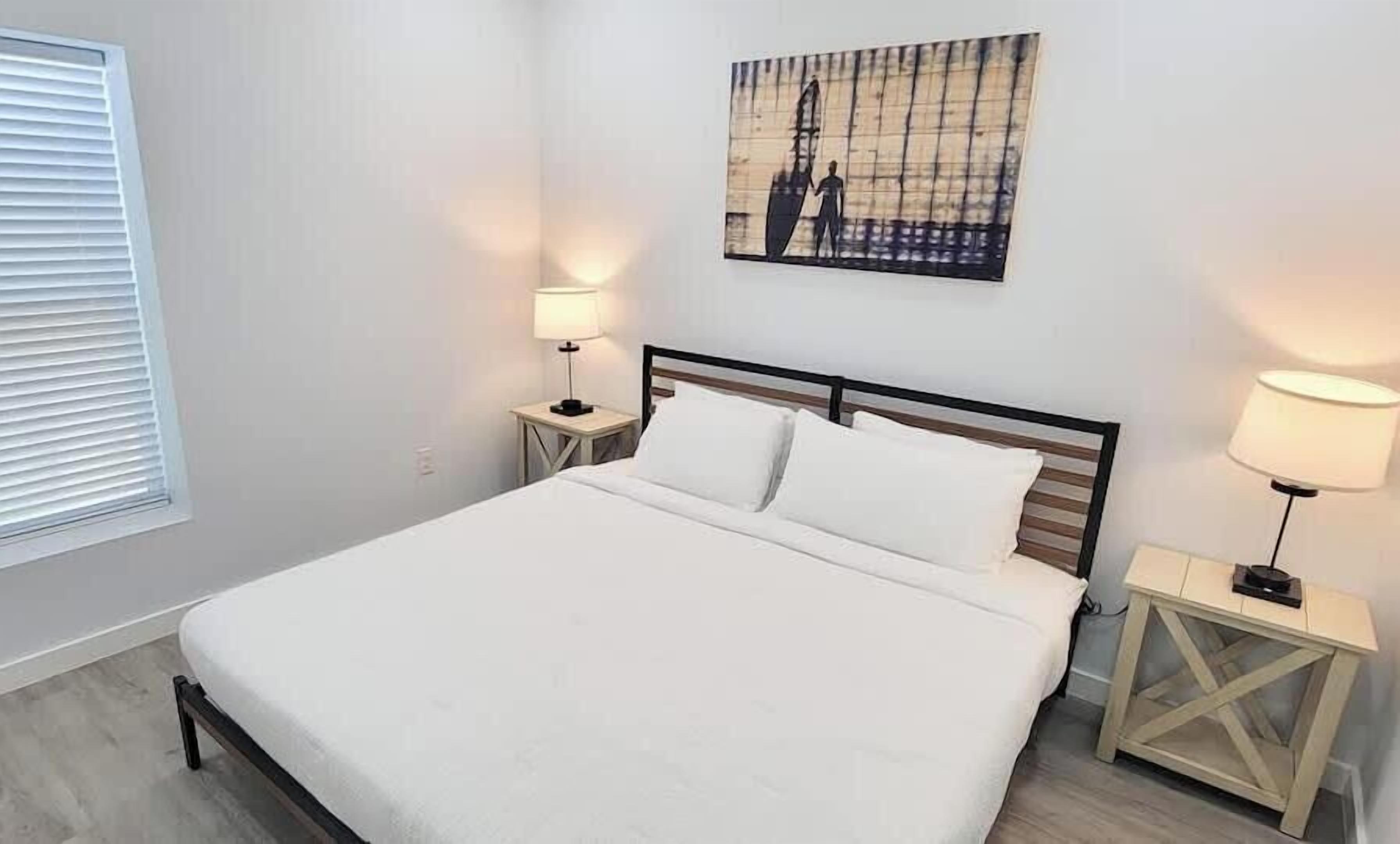 Un dormitorio con cama, dos mesitas de noche, una lámpara de pared, una ventana con persianas y un cuadro en la pared.