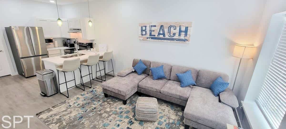 A modern living room with a grey sectional sofa, a small ottoman, and a patterned rug. There is a kitchen area with white cabinets and a dining table with chairs. A 'BEACH' sign is mounted on the wall.