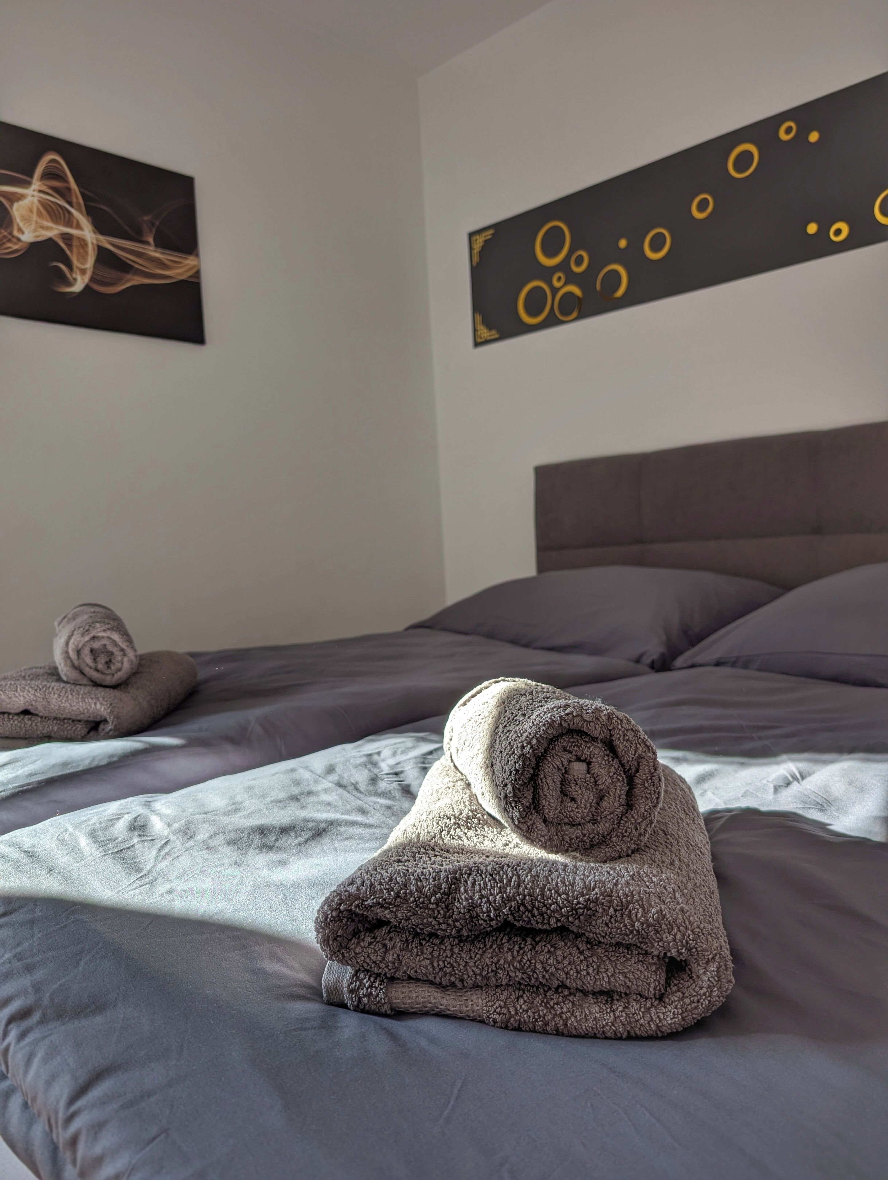 A neatly made bed with grey linens and a rolled-up towel on top. There are two wall art pieces, one with abstract designs and another with circular patterns.