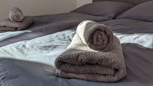 Ein ordentlich bezogenes Bett grauem Leinen und ein aufgerolltes Handtuch darauf. Zwei Wandbilder, eines mit abstrakten Mustern, das andere mit kreisförmigen Elementen.