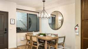 A dining area with a wooden table, chairs, a chandelier, a large mirror, and a window with blinds.