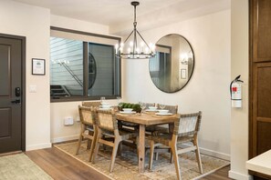 A dining area with a wooden table, chairs, a chandelier, a large mirror, and a window with blinds.