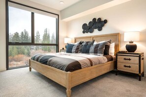 A bedroom with a large window, a bed with multiple pillows, a nightstand with a lamp, and a wall art piece.