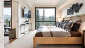 A bedroom with a bed, a TV, a window with a view, a wall art piece, and a door leading to a balcony.