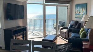 A living room with a TV, a sofa, a chair, and a table. There is a balcony with a view of the ocean.